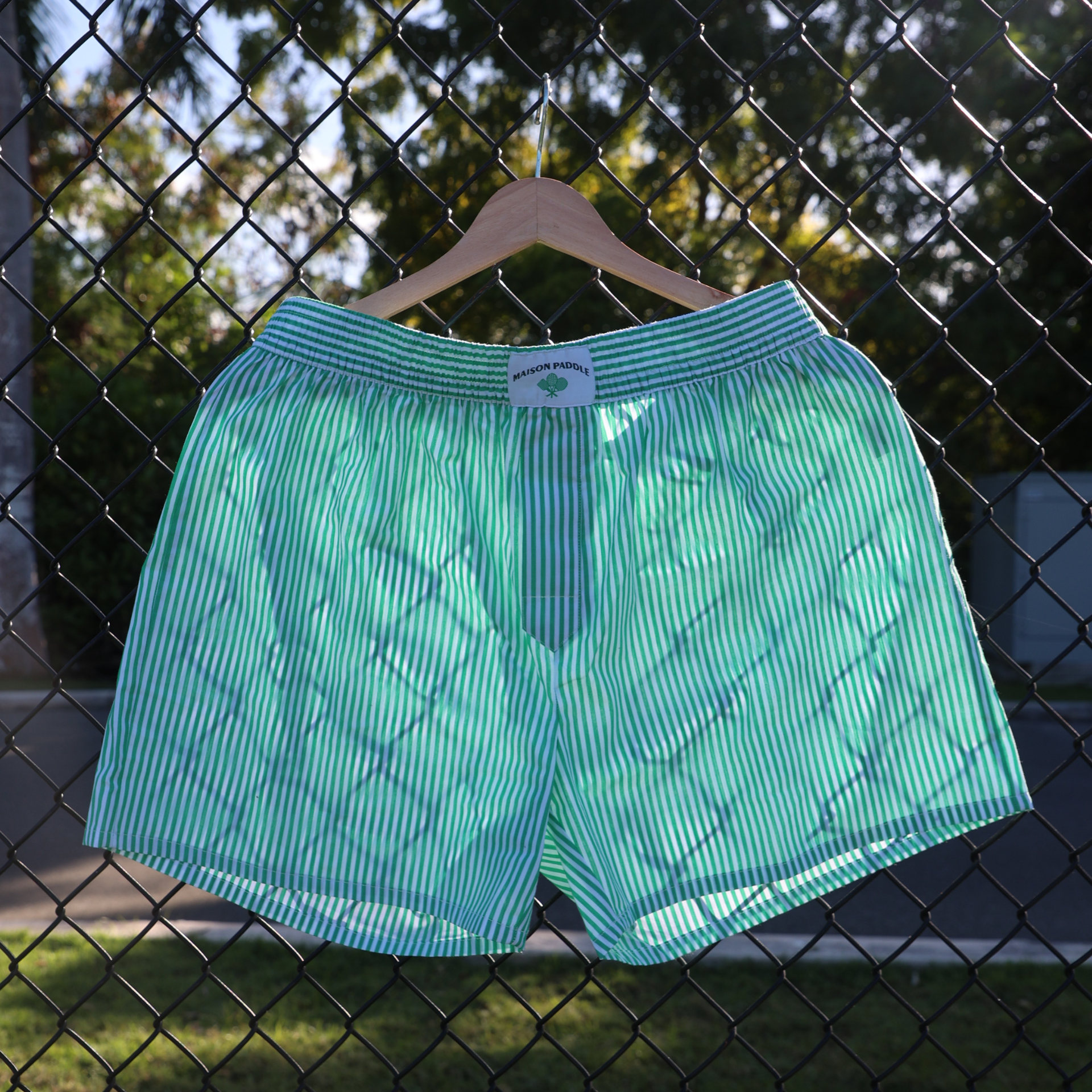 Maison Paddle striped boxer shorts hanging on a court fence, minimalist product shot for padel lifestyle apparel.